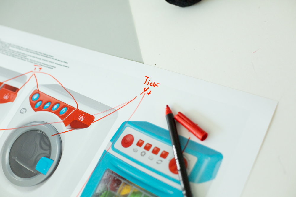 Close up von einem Ausdruck mit Layout für eine Spielzeug Waschmaschine, Korrekturen mit roten Linien, roter Stift liegt auf den Papier